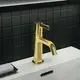 Goldene Waschtischarmatur von Ideal Standard mit einem gerade ausgeführten Auslauf und einem kompakten Bedienhebel, montiert auf einer weißen Oberfläche vor einer schwarzen Fliesenwand.